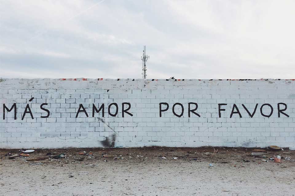 A photograph of a long wall with graffiti on. Text reads: Mas Amor Por Favor