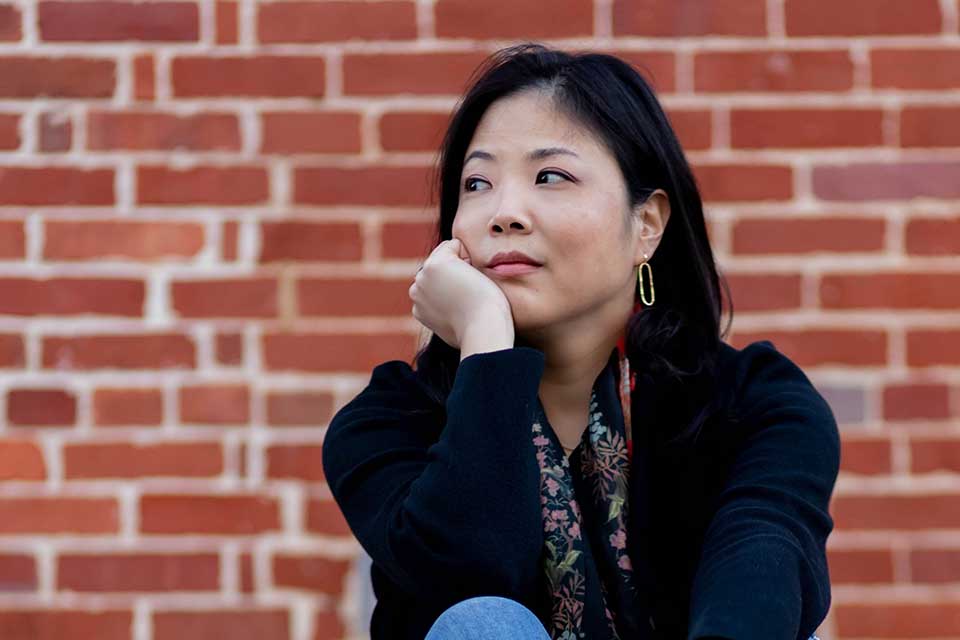 A photograph of Nicole Chung sitting in front of a red brick wall. She is looking off into the distance.